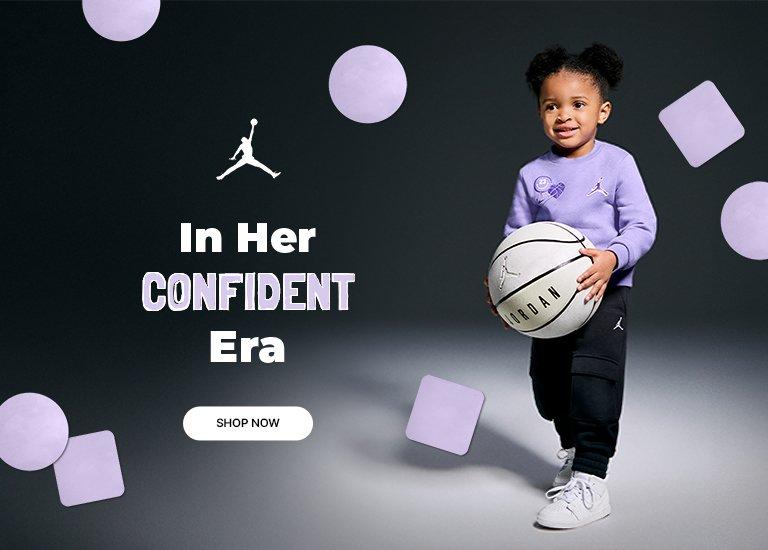 A split-image banner ad for Jordan apparel. On the left side, a little girl with her hair in two puffs smiles while holding a basketball. She is wearing a light purple crewneck sweatshirt with a Jordan logo and a heart graphic, black sweatpants, and white sneakers. The text "In Her CONFIDENT Era" is prominently displayed next to the Jordan "Jumpman" logo. On the right, a close-up of the same girl is smiling widely. Light purple circles and squares float around the images. A "SHOP NOW" button is at the bottom left.